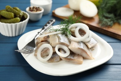 Plate with sliced salted herring fillet, onion rings and dill on blue wooden table, closeup