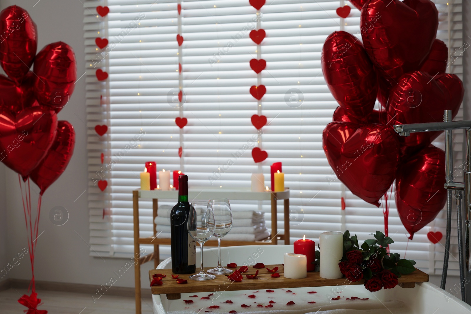 Photo of Wooden tray with wine, burning candles and rose petals on tub in bathroom. Valentine's day celebration