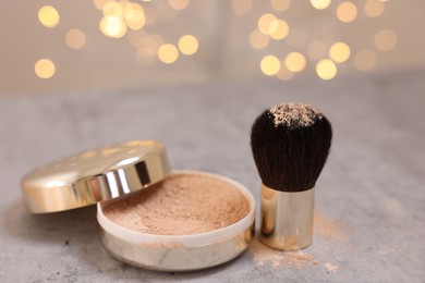 Face powder and brush on grey textured table against blurred lights, closeup