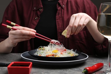 Photo of Stir-fry. Woman squeezing lime into tasty rice noodles with meat and vegetables at grey textured table, closeup