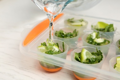 Photo of Pouring water into ice cube tray with lime slices and mint on table, closeup