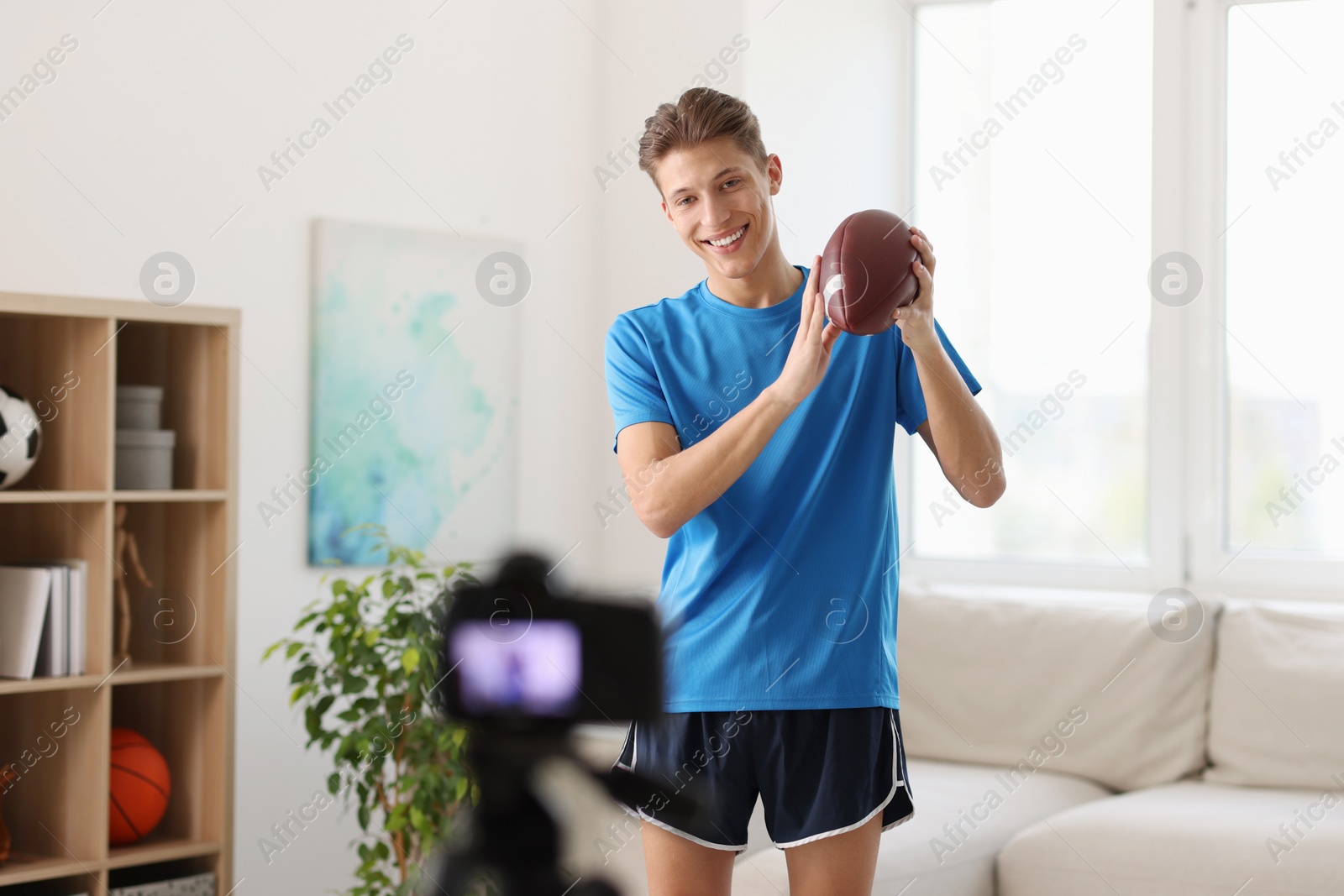 Photo of Smiling sports blogger holding american football ball while recording fitness lesson with camera at home