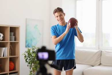 Smiling sports blogger holding american football ball while recording fitness lesson with camera at home