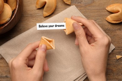 Image of Woman holding tasty fortune cookie with prediction Believe your dreams at wooden table, top view