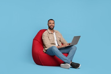 Smiling young man working with laptop on beanbag chair against light blue background