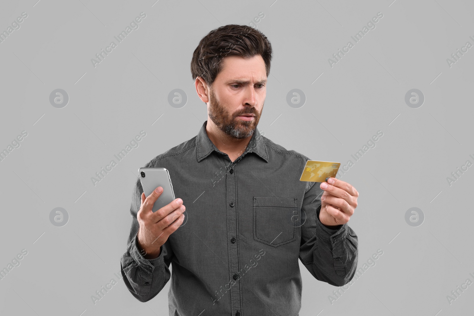 Photo of Emotional man holding smartphone and credit card on light grey background. Be careful - fraud