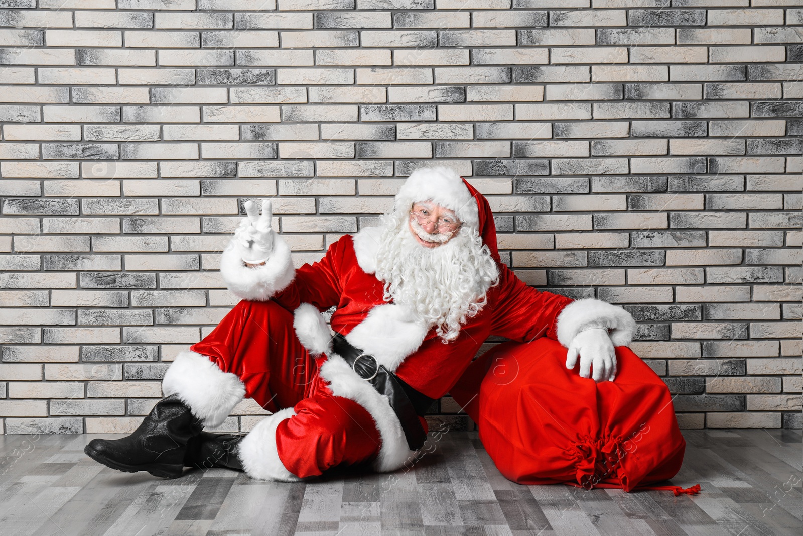 Photo of Authentic Santa Claus with big red bag full of gifts sitting on floor near brick wall