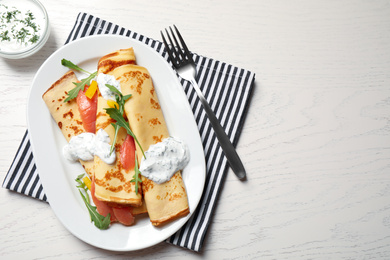 Delicious thin pancakes with salmon and sour cream on white wooden table, flat lay. Space for text