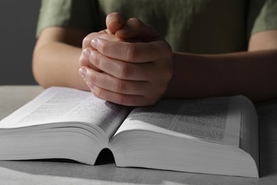 Woman holding hands clasped while praying over Bible at grey textured table, closeup