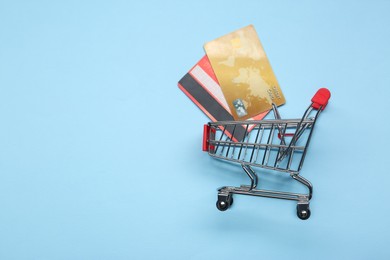Photo of Small metal shopping cart and credit cards on light blue background, top view. Space for text