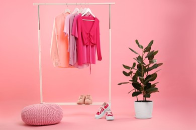 Rack with different stylish women`s clothes, sneakers and green plant on pink background