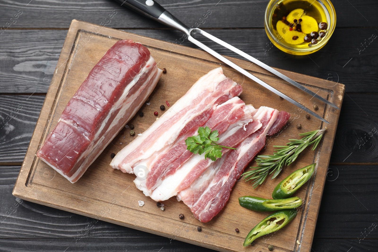 Photo of Pieces of raw pork belly, chili pepper, peppercorns, rosemary and parsley on black wooden table, flat lay