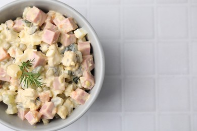 Tasty Olivier salad with boiled sausage in bowl on white tiled table, top view. Space for text