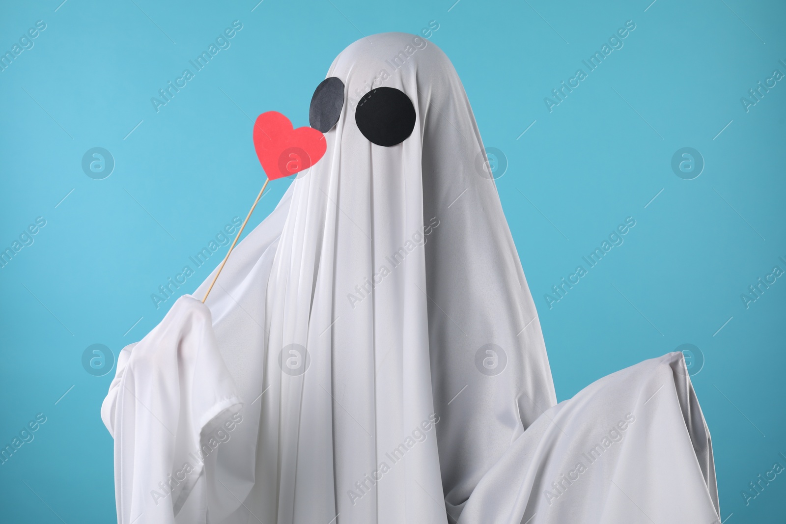 Photo of Cute ghost. Person covered with white sheet holding stick with red heart on light blue background