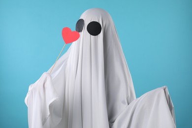 Cute ghost. Person covered with white sheet holding stick with red heart on light blue background