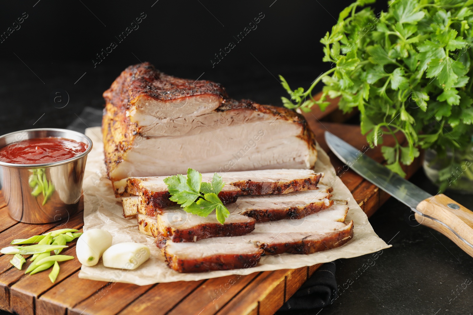 Photo of Pieces of baked pork belly served with sauce and parsley on black textured table, closeup