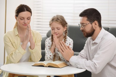 Girl and her godparents praying over Bible together at table indoors