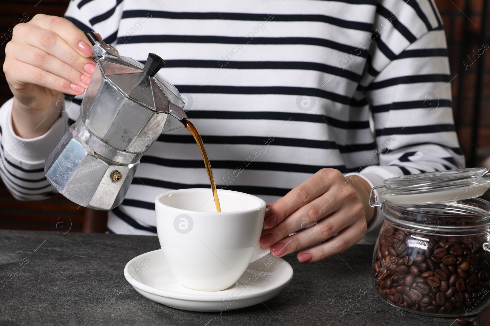 Photo of Woman pouring aromatic coffee from moka pot into cup at grey table indoors, closeup