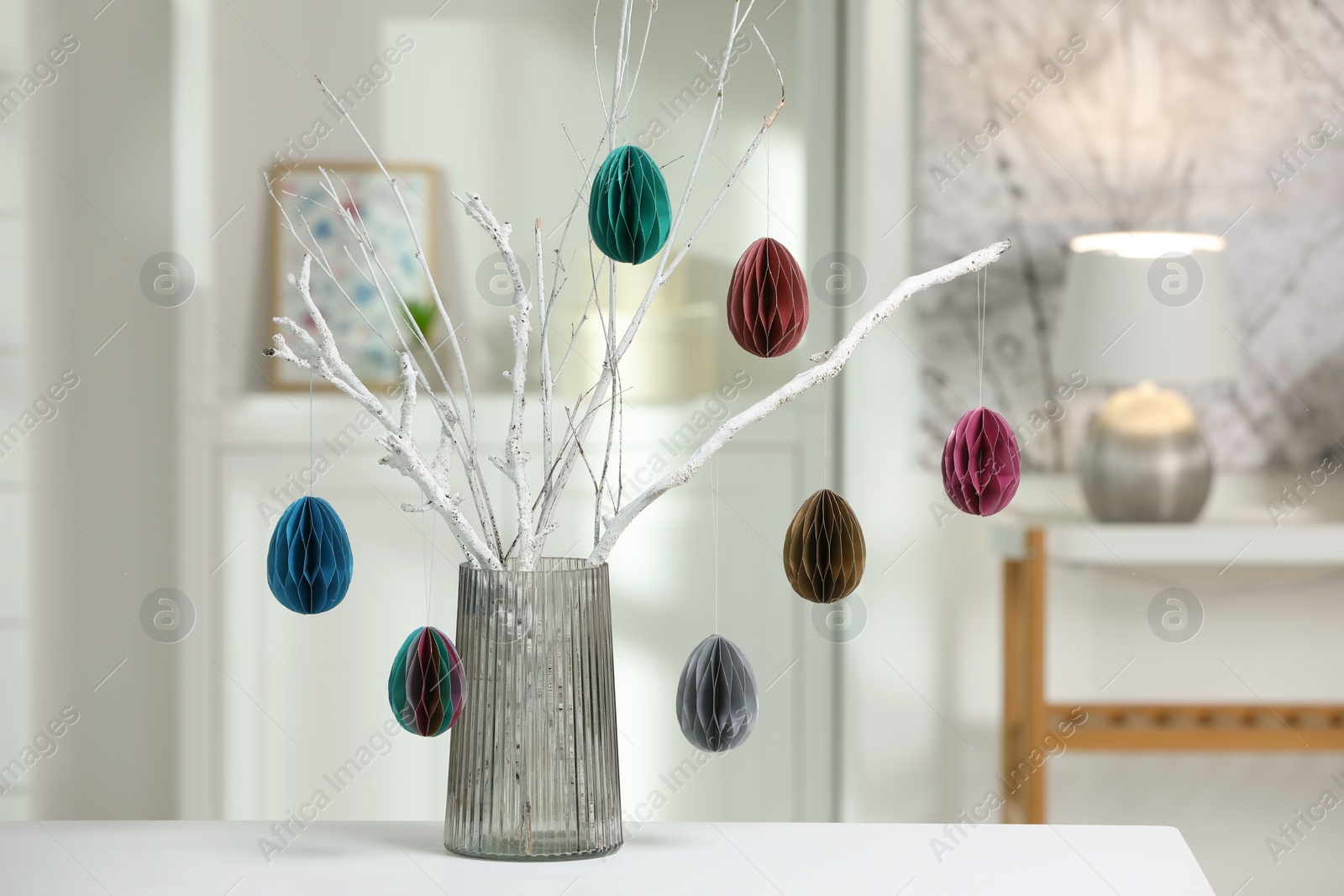 Photo of Branches with paper eggs in vase on white table at home. Beautiful Easter decor