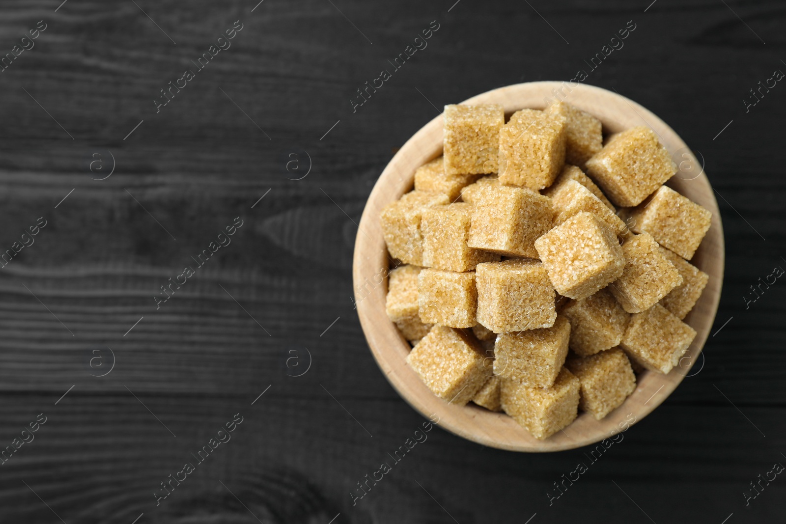 Photo of Brown sugar cubes in bowl on black wooden table, top view. Space for text