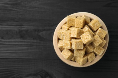 Photo of Brown sugar cubes in bowl on black wooden table, top view. Space for text