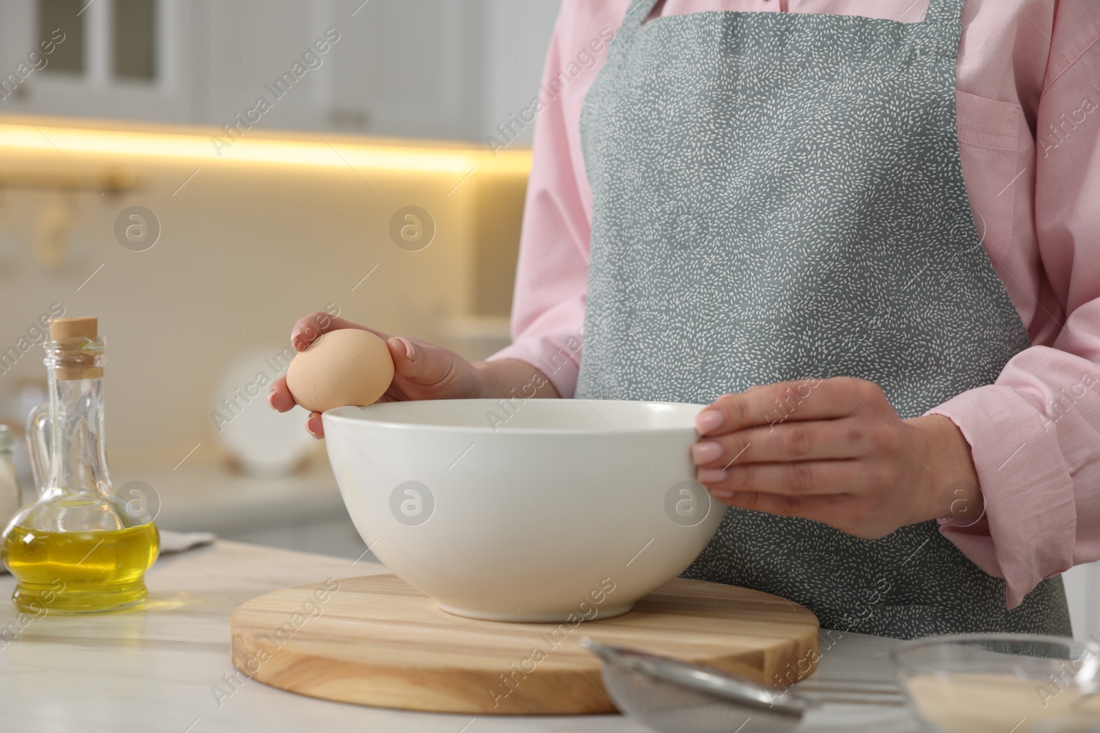 Photo of Making bread. Woman putting raw egg into bowl at white table in kitchen, closeup