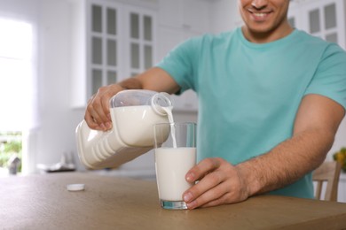 Man pouring milk from gallon bottle into glass at wooden table in kitchen, closeup
