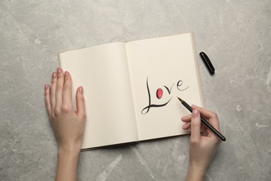 Woman writing word Love in notebook at light grey table, top view
