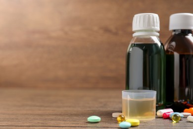 Bottles of syrup, measuring cup and pills on wooden table, space for text. Cold medicine