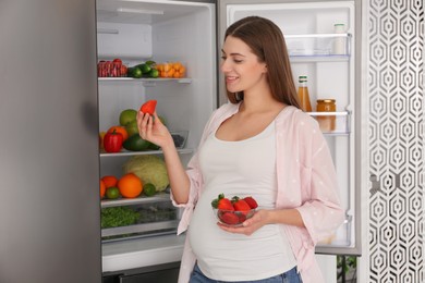 Photo of Young pregnant woman with strawberries near fridge at home. Healthy eating
