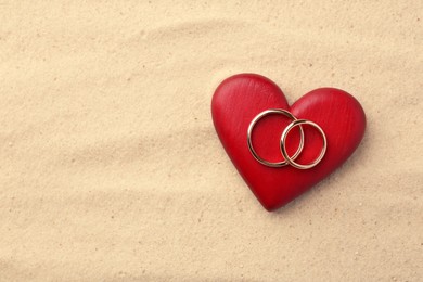 Honeymoon concept. Two golden rings and red heart on sand, top view. Space for text