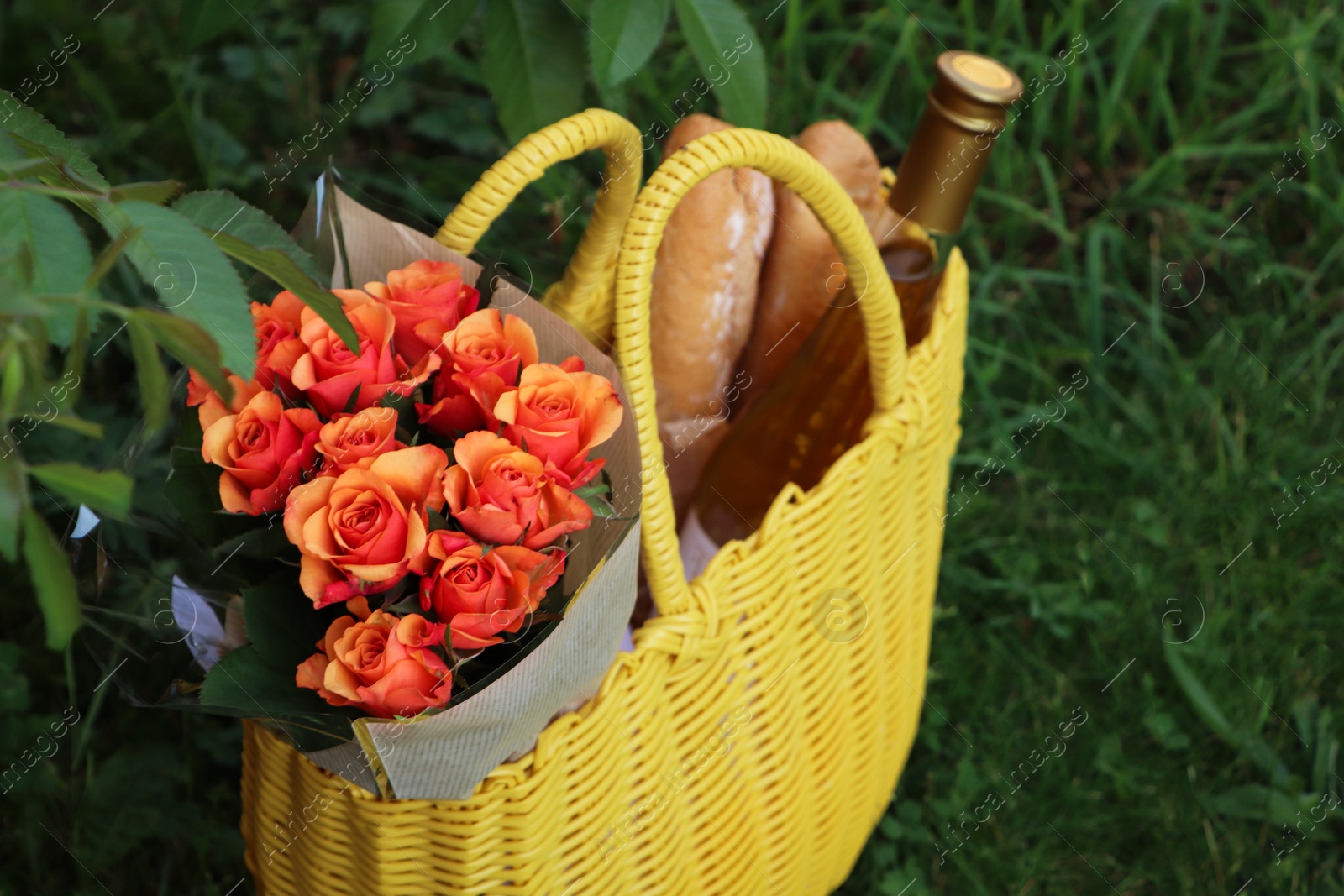 Photo of Yellow wicker bag with beautiful roses, bottle of wine and baguettes on green grass outdoors