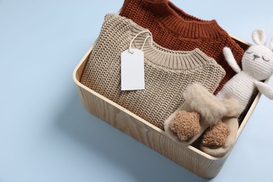 Photo of Different baby accessories, knitted sweaters and blank card in box on light blue background. Space for text