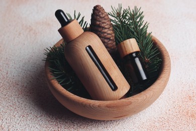 Photo of Bowl with bottles of aromatic essential oil, pine branches and cone on color table, closeup