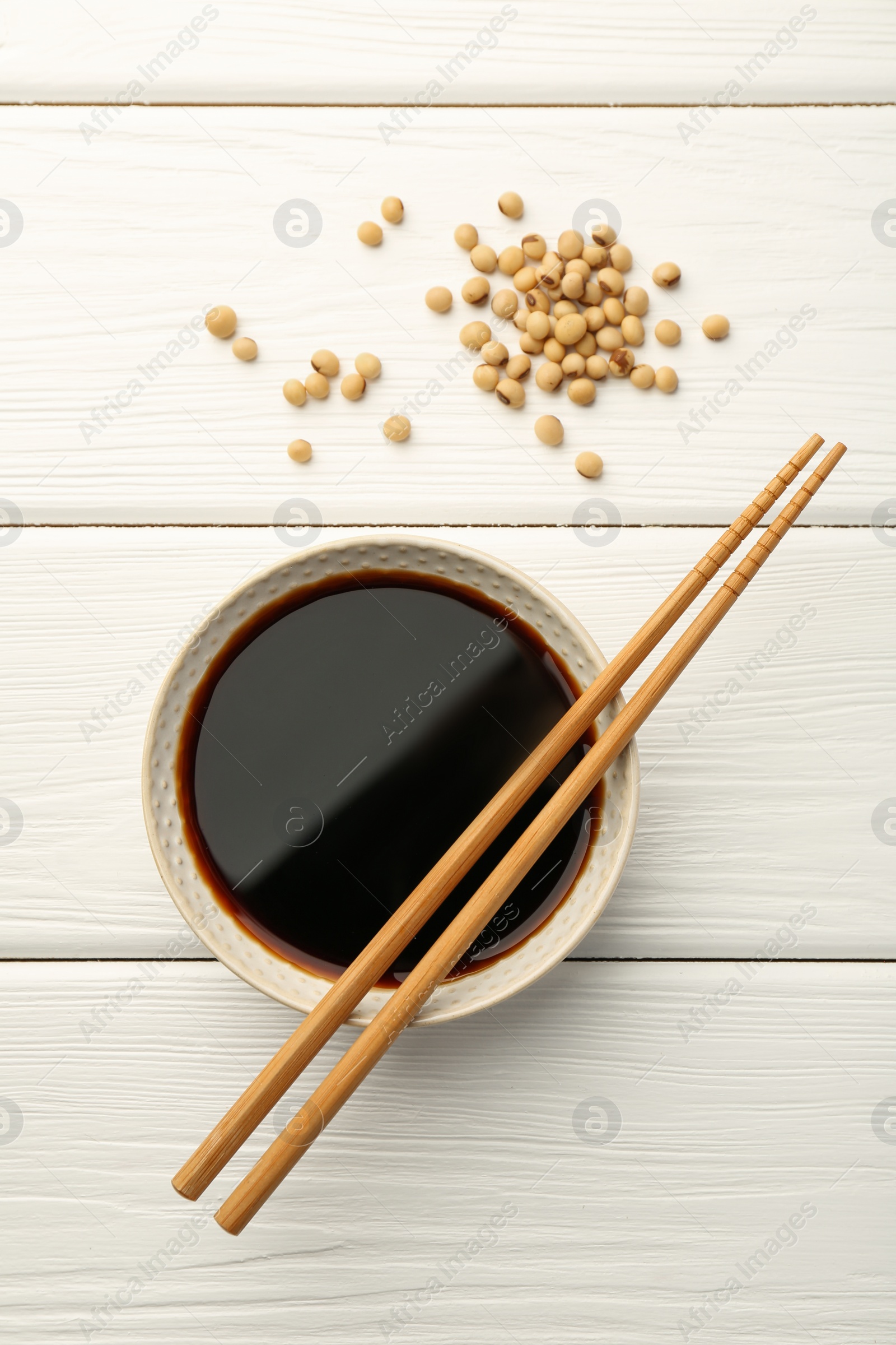 Photo of Tasty soy sauce in bowl, chopsticks and soybeans on white wooden table, top view