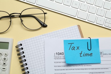 Reminder note with words Tax Time, notebook, computer keyboard, calculator and glasses on beige background, flat lay