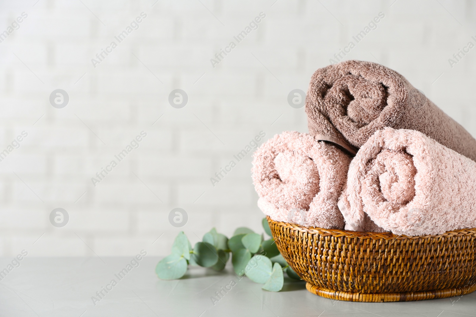 Photo of Wicker basket with rolled bath towels and green branch near white brick wall. Space for text