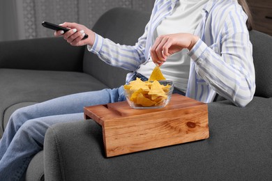 Woman holding remote control and eating nacho chips on sofa with wooden armrest table at home, closeup