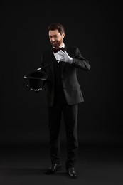 Happy magician showing magic trick with top hat on black background
