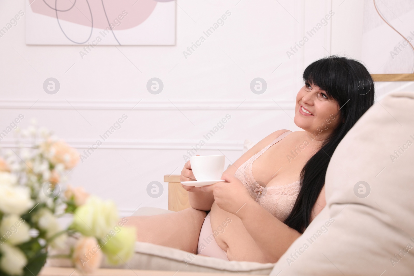 Photo of Beautiful overweight woman in beige underwear with cup of drink on sofa at home. Plus-size model