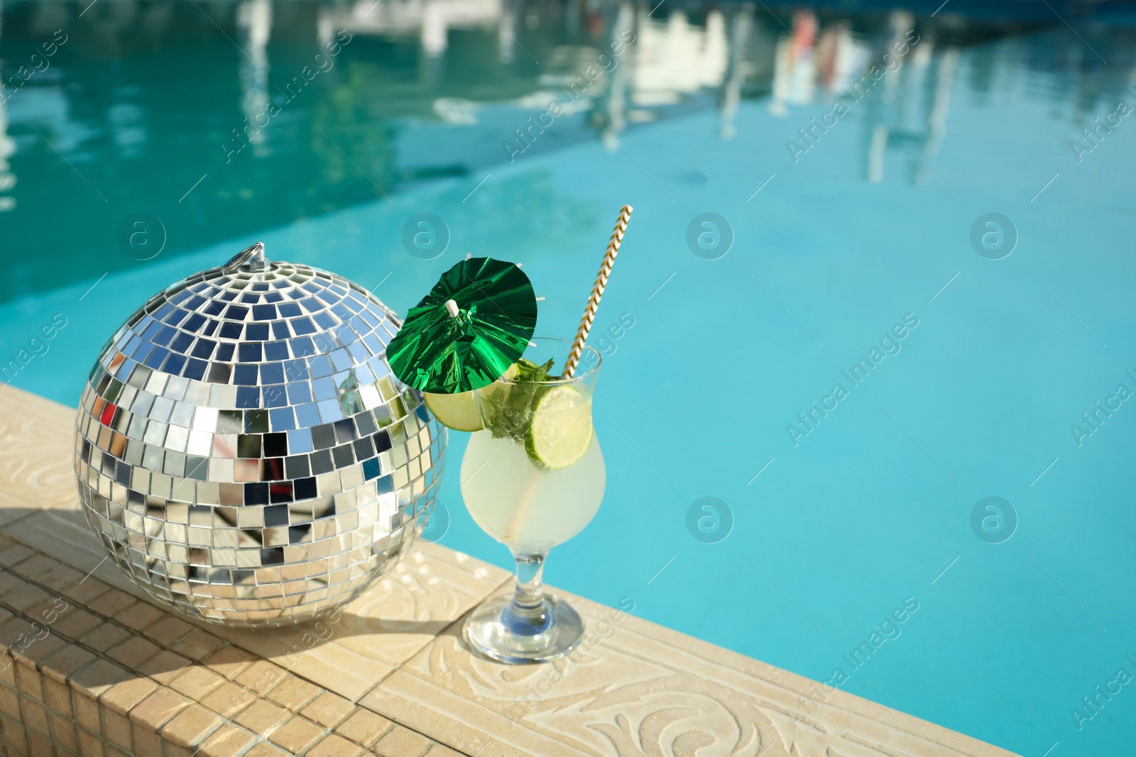 Photo of Tasty refreshing cocktail and shiny disco ball on edge of swimming pool. Party items