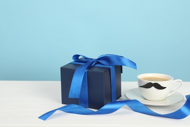 Photo of Happy Father's Day. Cup with moustache and gift box on white wooden table, closeup. Space for text