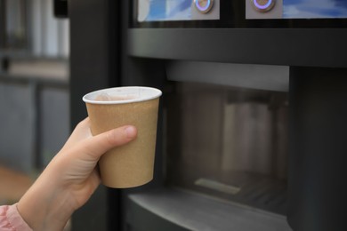Photo of Girl holding paper cup with drink near coffee and hot beverage vending machine, closeup. Space for text