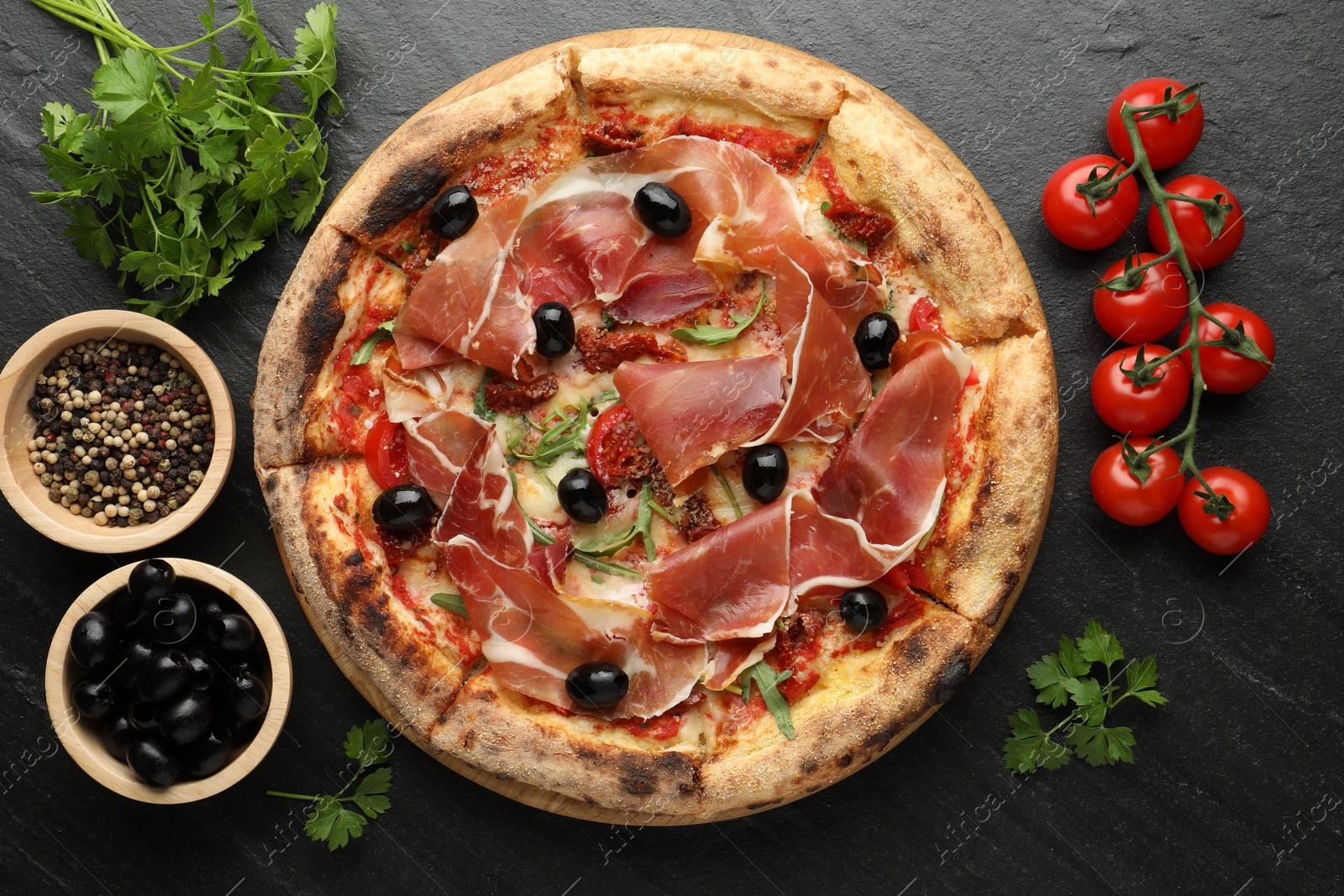 Photo of Tasty pizza with cured ham, olives, tomatoes, parsley and spices on black table, top view