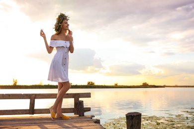 Young woman wearing wreath made of beautiful flowers on pier near river