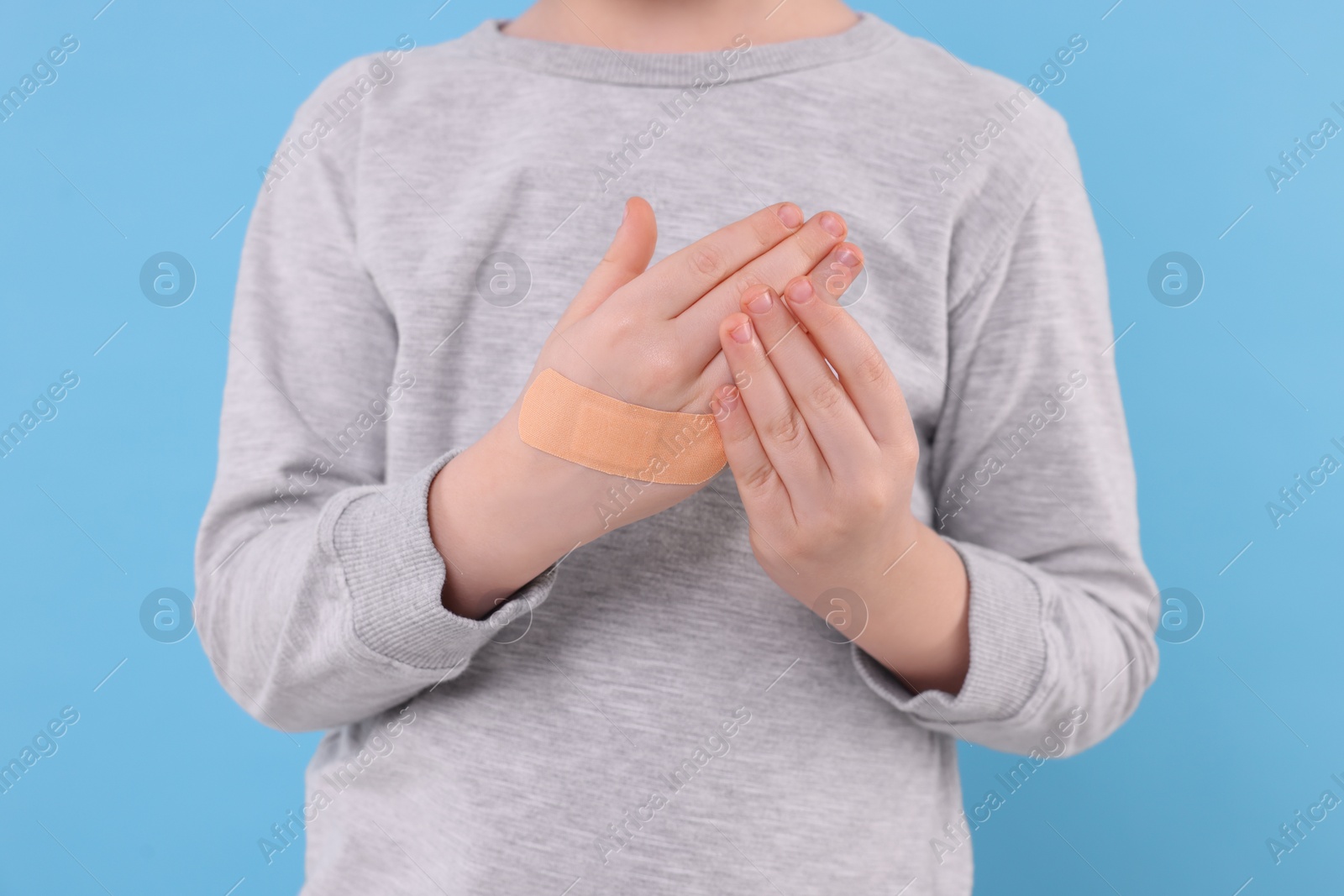 Photo of Little boy putting sticking plaster onto hand on light blue background, closeup