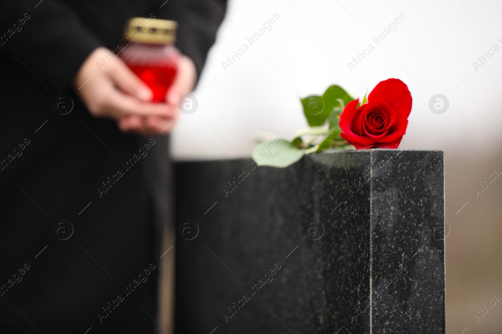 Photo of Woman with candle near black granite tombstone outdoors, focus on red rose. Funeral ceremony