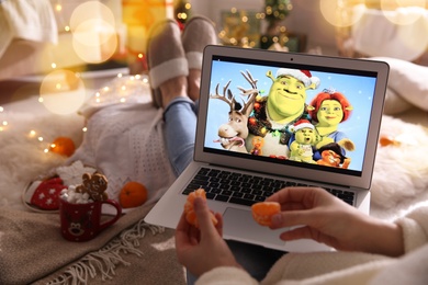 MYKOLAIV, UKRAINE - DECEMBER 25, 2020: Woman with tangerine watching Shrek The Halls movie on laptop at home, closeup. Cozy winter holidays atmosphere
