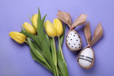 Easter bunnies made of craft paper and eggs near beautiful tulips on purple background, flat lay