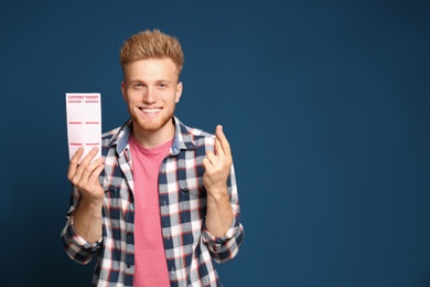 Portrait of hopeful young man with crossed fingers holding lottery ticket on blue background